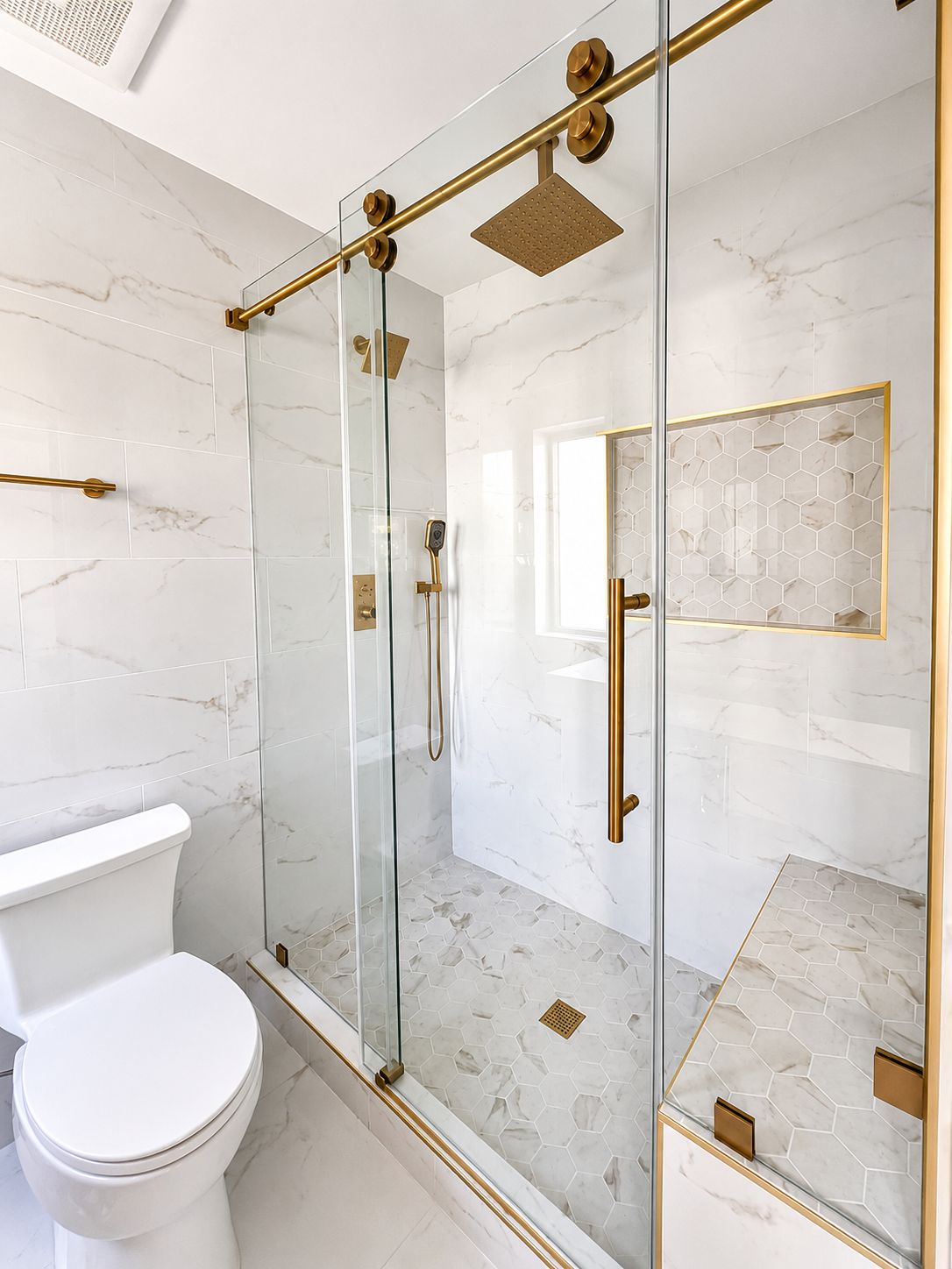 Glass-enclosed marble shower with gold hardware and hexagon tile bench